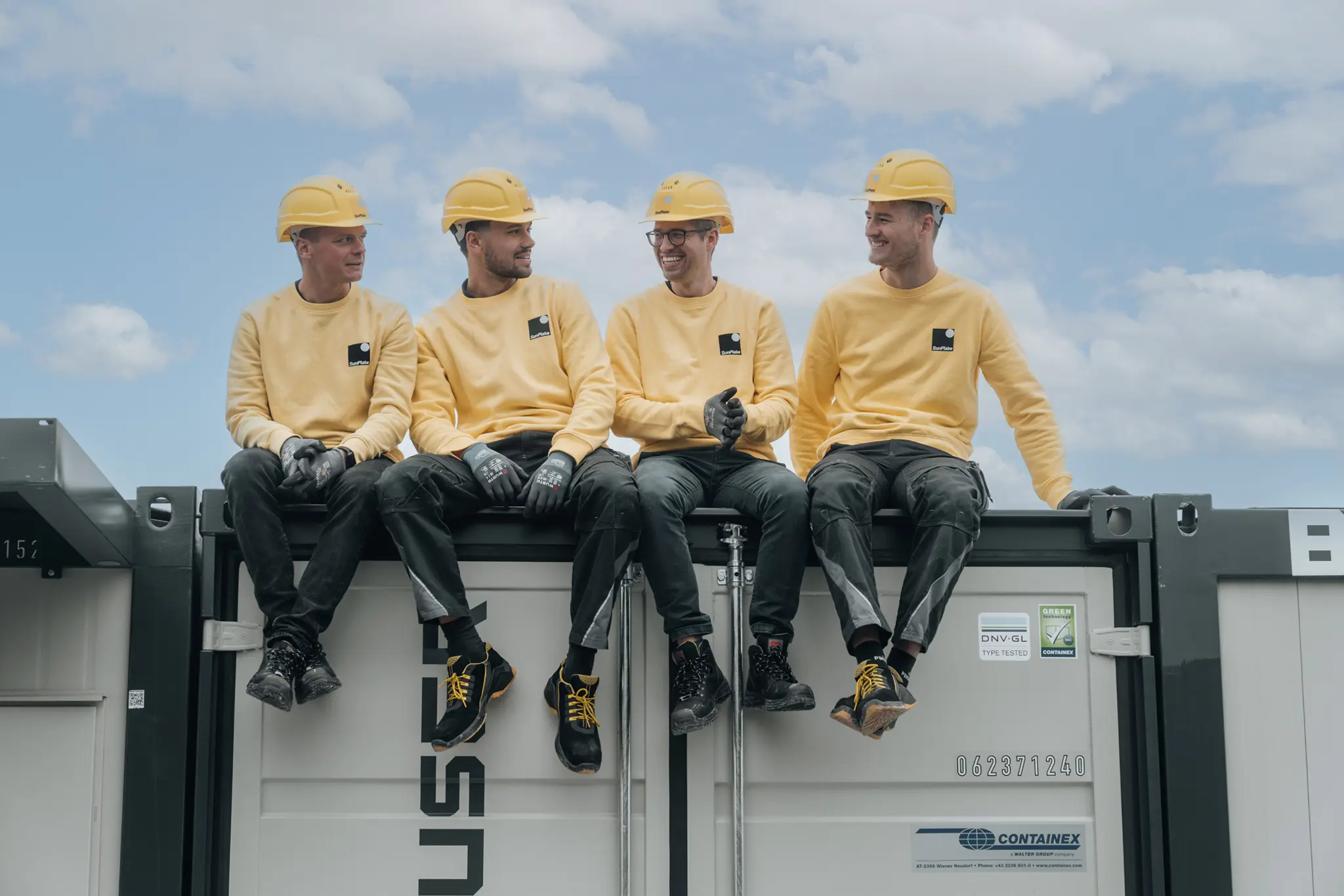 Vier Bauarbeiter in gelben Helmen und Arbeitskleidung sitzen lachend nebeneinander auf einem Container unter blauem, leicht bewölktem Himmel.