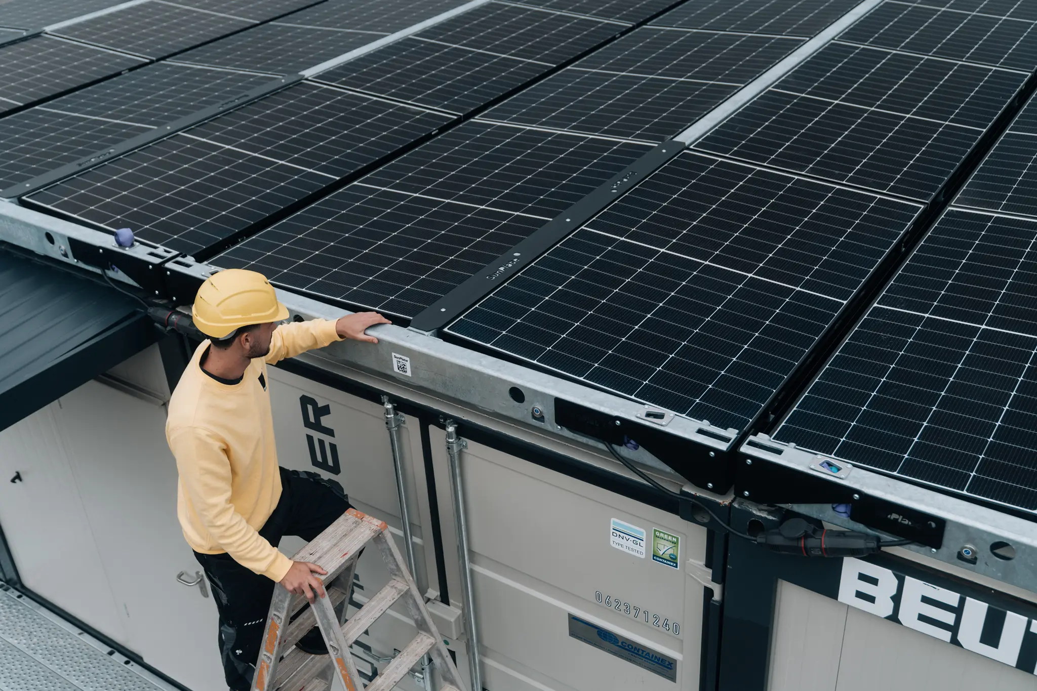 Ein Arbeiter mit gelbem Schutzhelm steht auf einer Leiter und kontrolliert die Solarmodule auf dem Dach eines Containers.